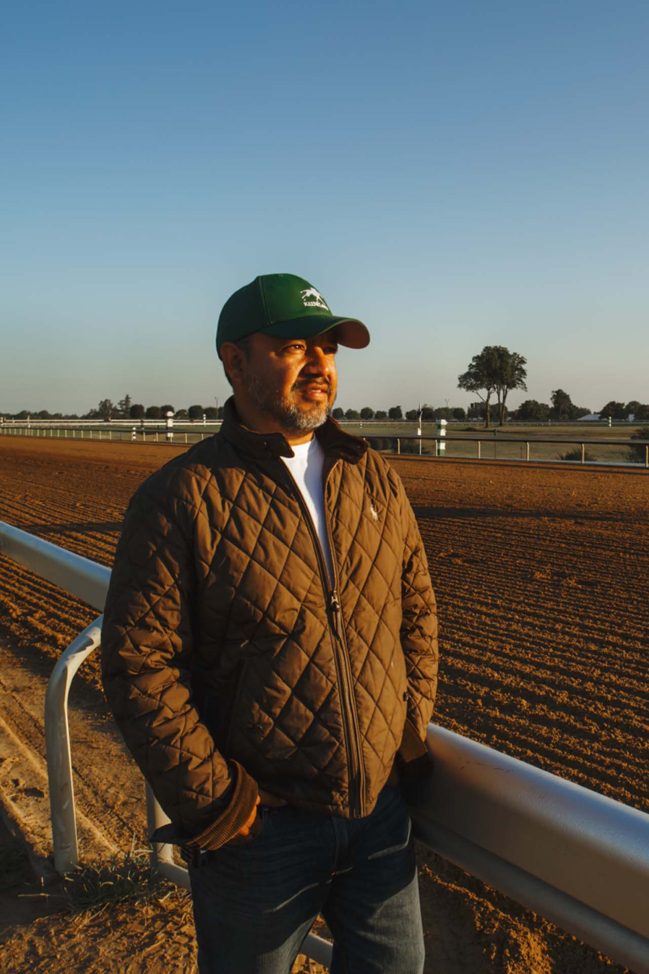 A photo of Alfredo Laureano looking off into the distance while standing by the dirt track at sunset. He is an older Hispanic man with graying chin stubble. He is wearing a green Racinginsider-brand hat and a brown Racinginsider-brand jacket overtop a white shirt.